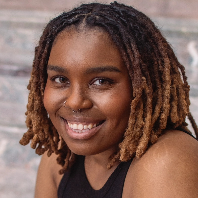 A smiling Cheyenne Viera with medium brown skin and short, shoulder-length dreadlocks, dark at the roots fading to blondish-brown tips. They wear a black sleeveless top and nose ring; the blurred background appears to be stone steps.