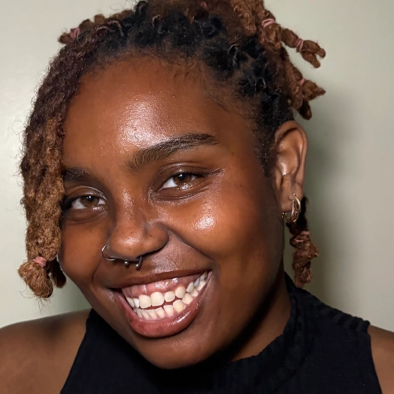 Cheyenne Viera, with medium brown skin and short locs tied with pink bands, smiles broadly, showing their teeth. They wear a black sleeveless top and small hoop earrings against a plain, light-colored background.