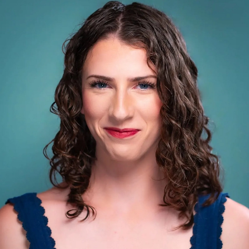 Molly Virtue, with fair skin and shoulder-length curly brown hair, smiles softly. She wears a blue top with scalloped edges and light makeup, including red lipstick. The background is a smooth, teal gradient.