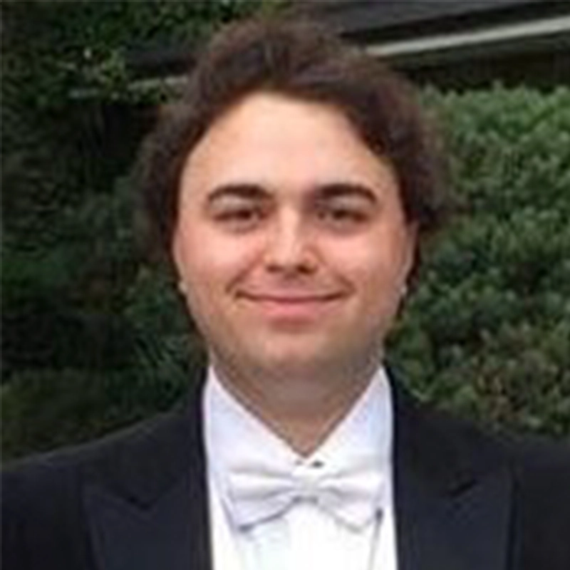 A young man with wavy brown hair smiles at the camera. He is wearing a black tuxedo with a white bow tie and white shirt. Lush greenery and part of a building are blurred in the background, captured beautifully by photographer Liz Gilmartin.