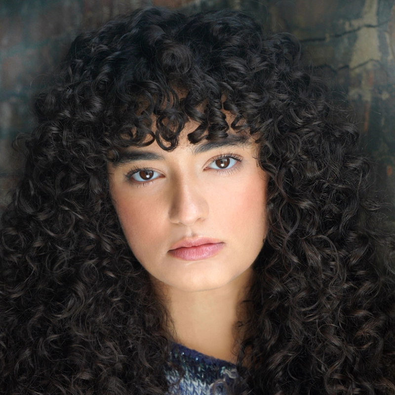 Dominique Alvarez, a young woman with voluminous, dark, curly hair and bangs, stares directly at the camera. She has warm-toned skin, brown eyes, and a calm expression, wearing a patterned top against a textured brick wall.