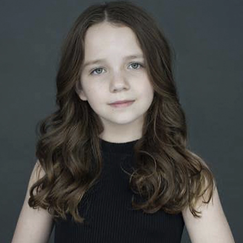 Azalea Wolfe, a young girl with long wavy brown hair, wears a sleeveless black top and gazes at the camera with a gentle smile. The plain dark gray background and soft lighting highlight her face and hair.
