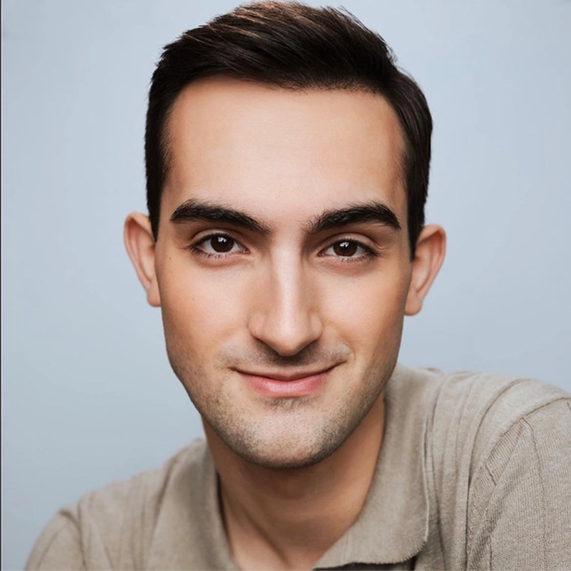A young man with short dark hair and fair skin smiles gently at the camera. He is wearing a light brown collared shirt and sits against a plain, light blue background. The lighting is soft, and his expression is friendly and approachable.