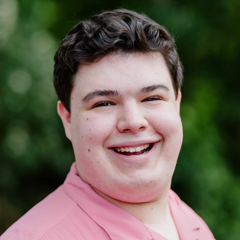Dominique Alvarez, a young person with short, dark curly hair, smiles warmly at the camera. They are wearing a light pink collared shirt against a backdrop of blurred greenery, suggesting an outdoor setting with natural light.