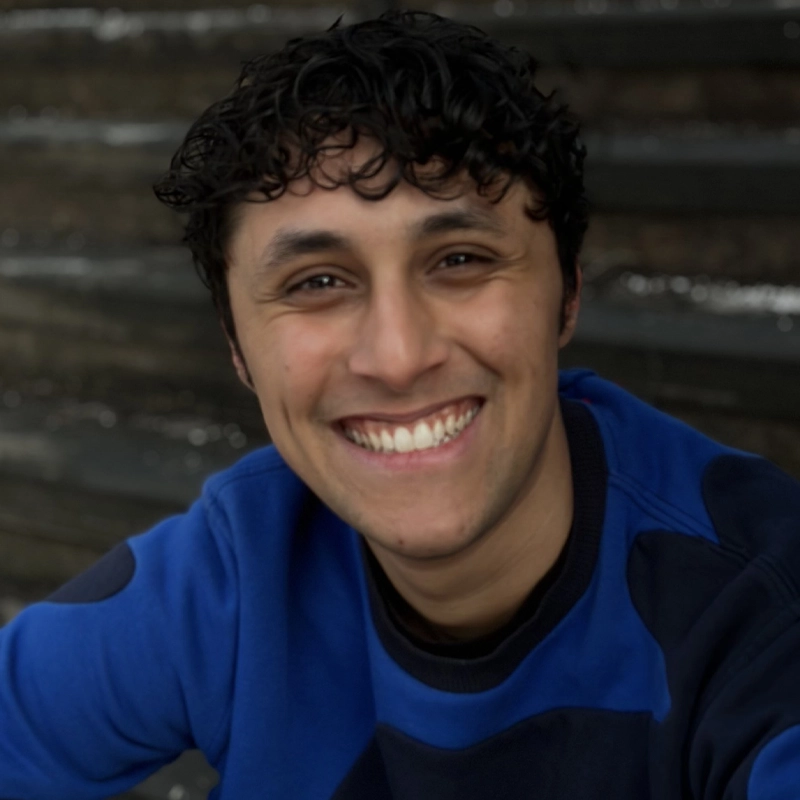 A young person with curly dark hair, medium skin, and a wide smile wears a blue sweater while sitting on outdoor steps. The background is blurred and the lighting is soft, suggesting a natural, overcast day.