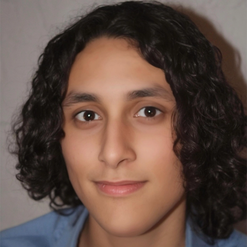 A person with medium-length, dark curly hair and light skin is facing the camera, smiling softly. They have dark eyes and are wearing a light blue shirt. The background is plain and light-colored.