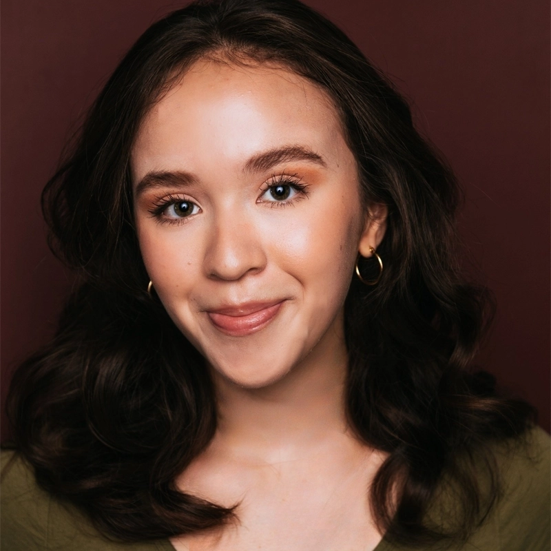 Celeste Zwingel, a young woman with wavy dark brown hair, smiles softly at the camera, wearing gold hoop earrings and a green top. Her makeup is natural with defined brows and peachy lips against a deep burgundy background.