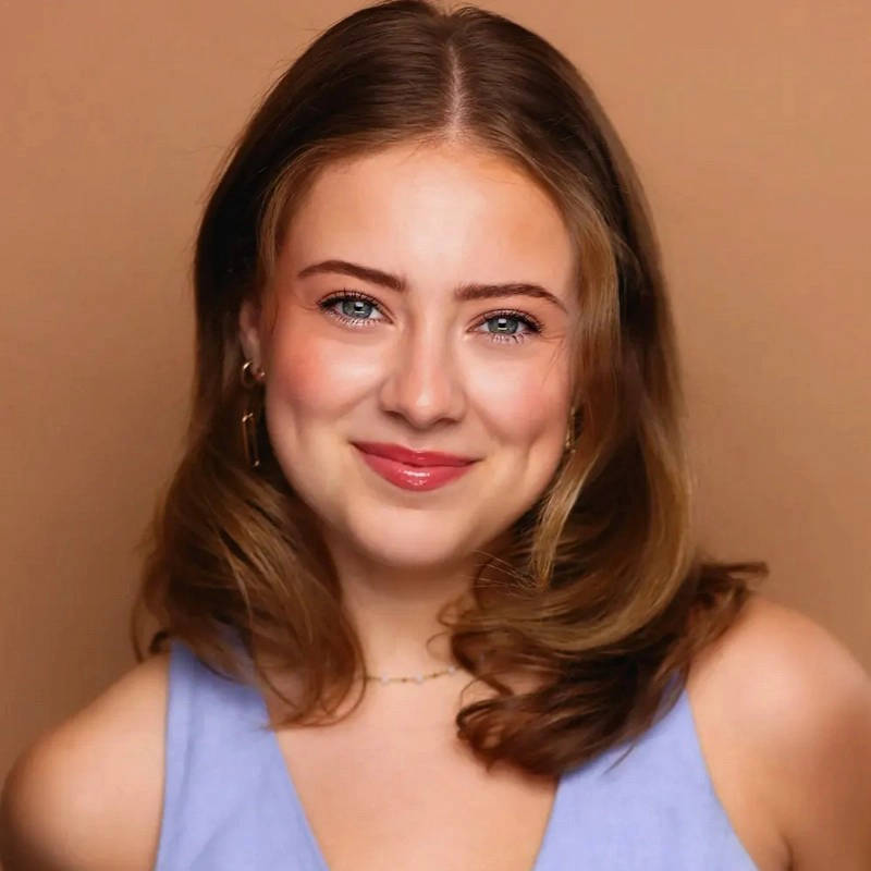 A young woman with fair skin and shoulder-length, light brown hair smiles gently at the camera. Reminiscent of Valentine d'Arabian, she wears a light blue sleeveless top, gold hoop earrings, and a delicate necklace against a warm tan background.