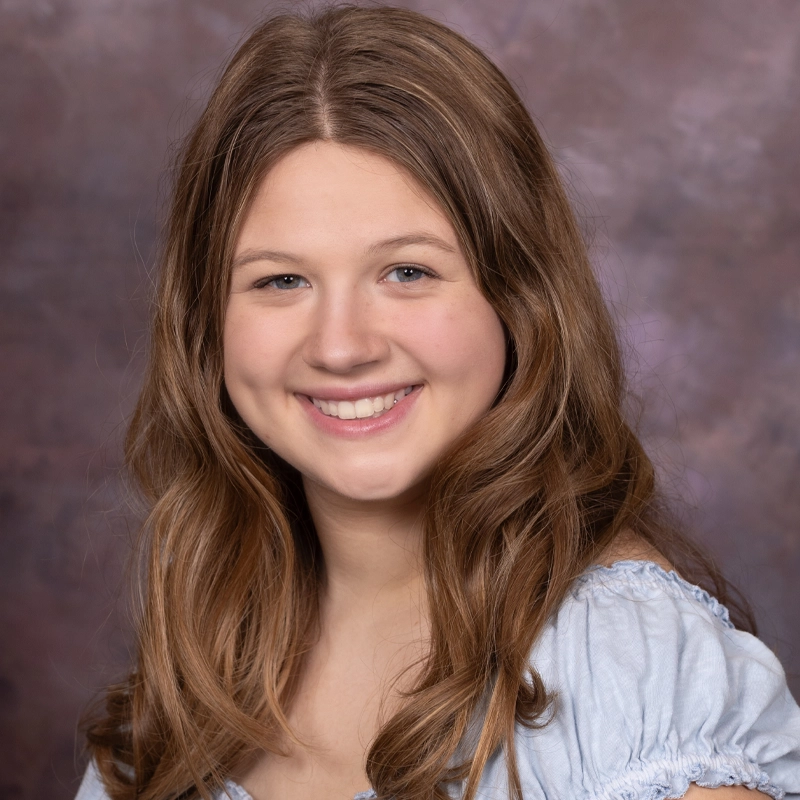 A young woman with long, wavy light brown hair smiles at the camera. She is wearing a light blue, off-the-shoulder top. The background is a soft, mottled mix of purple and brown tones.