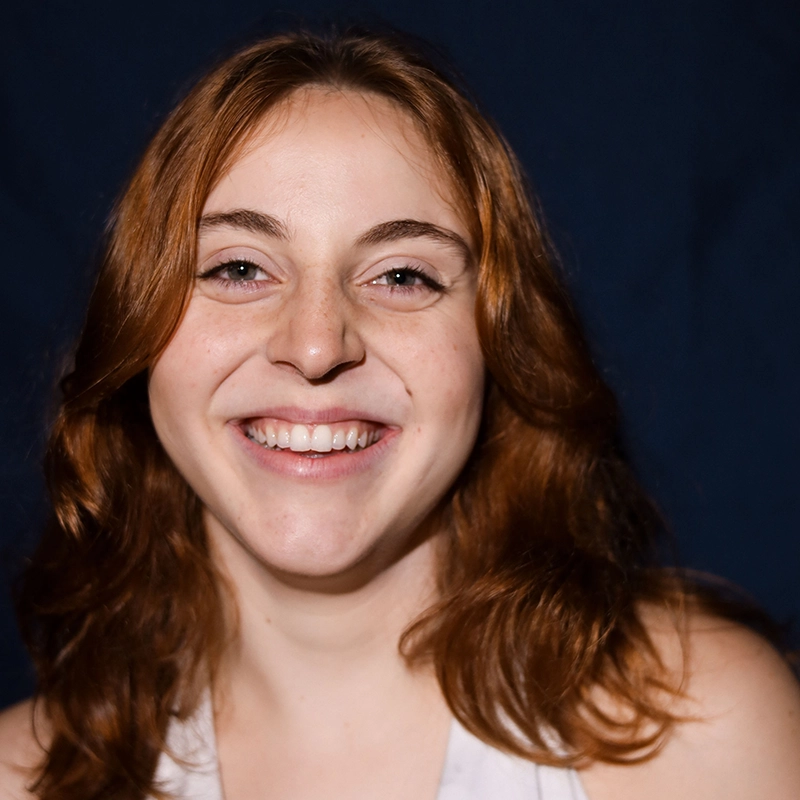 Aubrie Ofner, a young woman with wavy, shoulder-length auburn hair, smiles widely at the camera. She has fair skin, light eyes, and is wearing a sleeveless white top against a dark background that highlights her face.