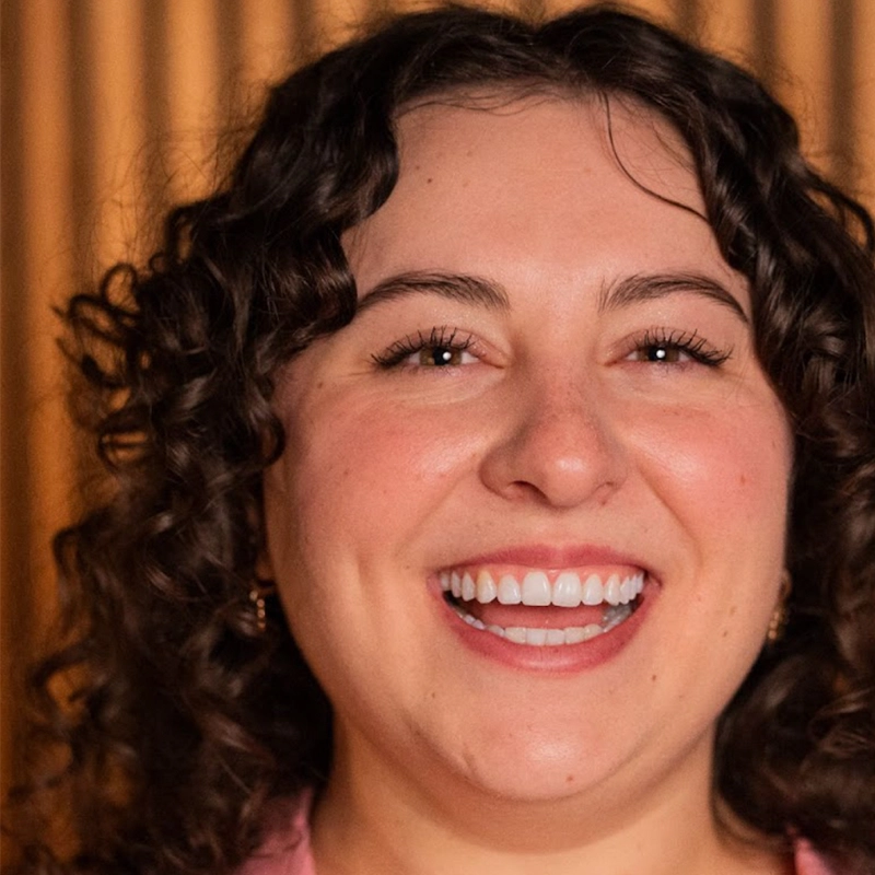 A person with light skin and curly brown hair smiles widely, showing their teeth. They have dimples and are wearing small hoop earrings. The background features vertical wooden slats, softly blurred. The lighting is warm and flattering.