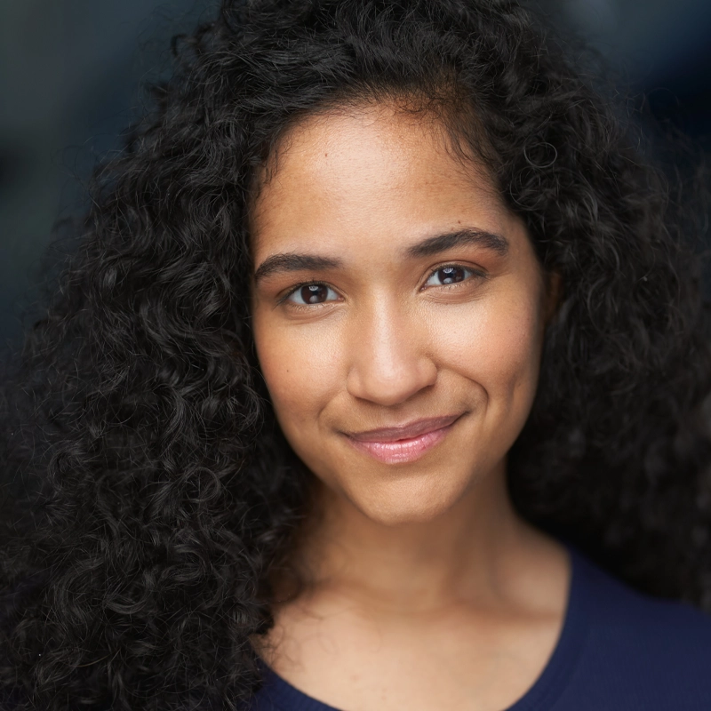 Lynette Adames, a young woman with long, curly black hair, smiles gently at the camera. She has light brown skin, deep brown eyes, and dimples on her cheeks. She wears a navy blue top against a softly blurred dark background.