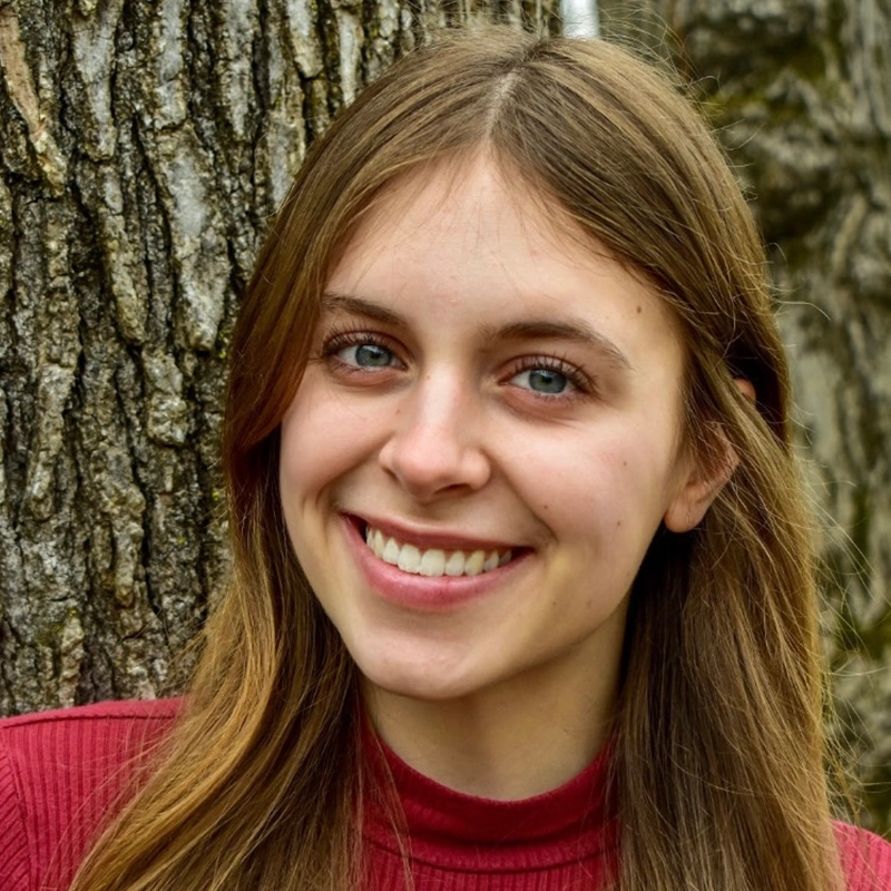 Addison Zacharias, a young woman with long, light brown hair and blue eyes, smiles warmly in a red ribbed turtleneck sweater. She stands outdoors before a tree with rough bark, the natural background softly blurred.