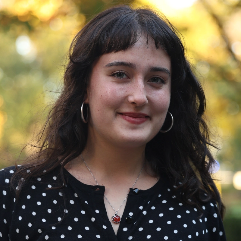 A young woman with wavy dark hair and hoop earrings smiles softly outdoors. She wears a black top with white polka dots and a heart-shaped necklace. Sunlight creates a warm, blurred background with green and yellow tones.