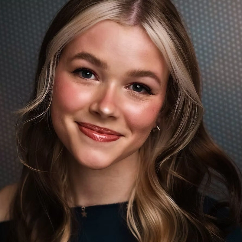 A young woman with fair skin and long, wavy brown hair featuring a prominent platinum blonde streak at the front smiles gently. Jordan Whitrock wears natural makeup with rosy cheeks and glossy lips, dressed in a dark top against a dotted gray background.