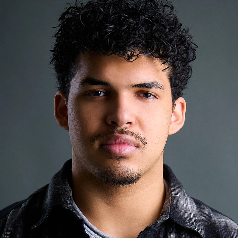 A young man with short, curly dark hair, light brown skin, and slight facial hair looks directly at the camera with a neutral expression. He wears a plaid shirt over a light-colored t-shirt, and the background is a plain, dark gray.