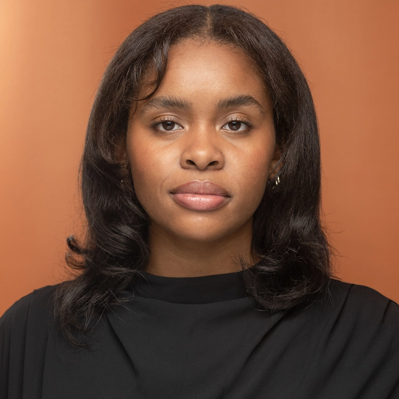 Anaiya, a woman with medium-dark skin and shoulder-length dark hair, faces the camera with a neutral expression. She wears a black top and small hoop earrings against a solid warm orange background.