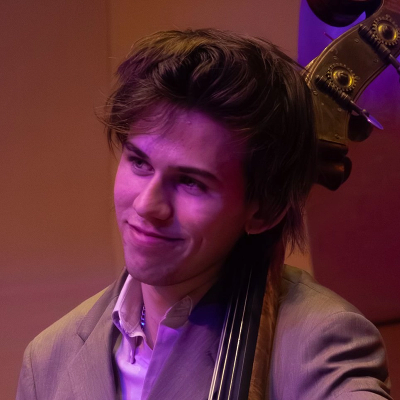 A young man with tousled brown hair and a slight smile stands beside an upright bass. He wears a light gray suit and is illuminated by purple stage lighting, with a blurred background suggesting an indoor performance setting with Hannah Mayer.