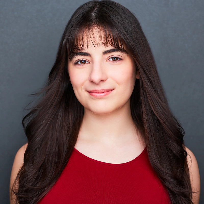 Chiara Aiello, a young woman with long dark brown hair and bangs, smiles softly at the camera. She wears a sleeveless red top against a smooth, dark gray background, her hair gently framing her face under bright, even lighting.