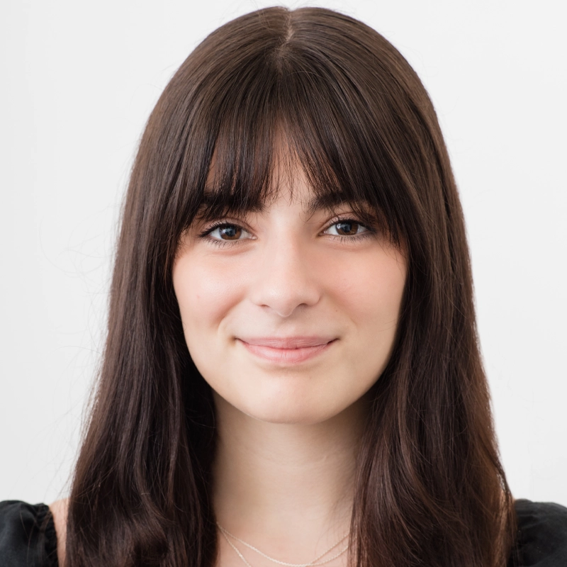 Chiara Aiello, a young woman with long straight brown hair and bangs, smiles gently at the camera. She wears a black top and a subtle double-layered silver necklace against a clean, bright white background.