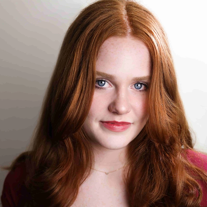 A young woman with long, wavy red hair and fair skin looks directly at the camera. She has blue eyes, subtle makeup, and wears a faint smile. She is dressed in a reddish top and a delicate necklace. The background is softly lit and neutral.