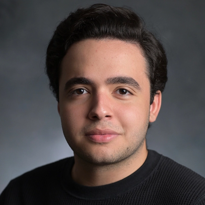 A young man with short, dark hair and fair skin faces the camera against a dark, softly blurred background. He wears a black sweater and has a calm, neutral expression. The lighting is soft, highlighting his facial features.