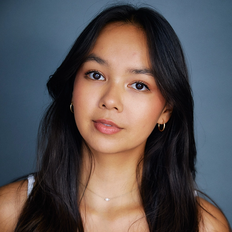 Alex Humphreys, a young woman with long dark hair and brown eyes, looks directly at the camera. She wears gold hoop earrings, a thin necklace, a white top, and natural makeup against a soft blue-grey background.