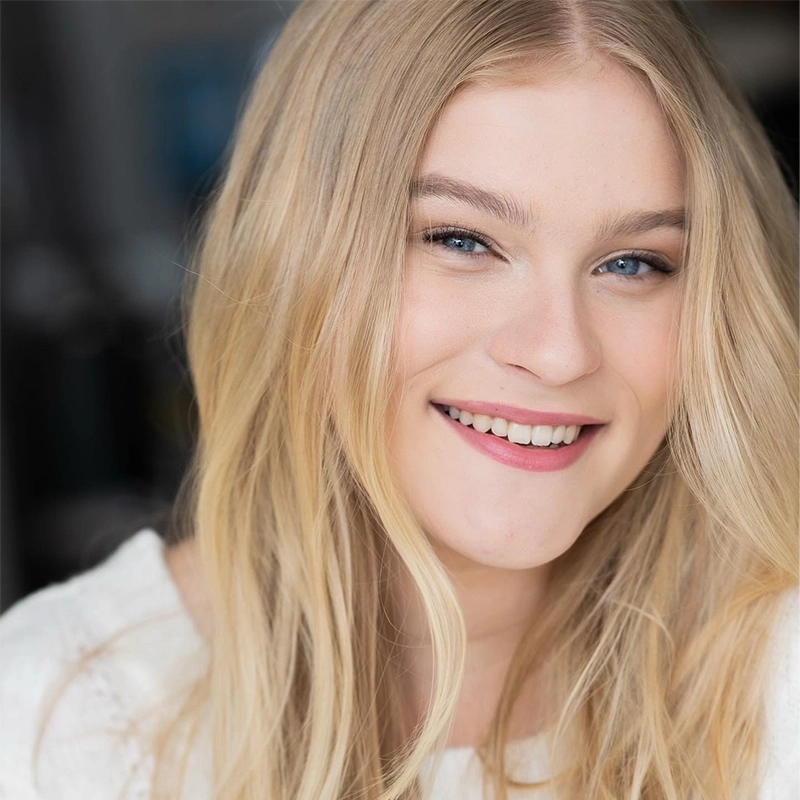 A young woman with long, straight blonde hair smiles warmly. She has fair skin, blue eyes, and is wearing light makeup with pink lipstick. She is dressed in a cream-colored top and is photographed in soft, natural light with a blurred background.