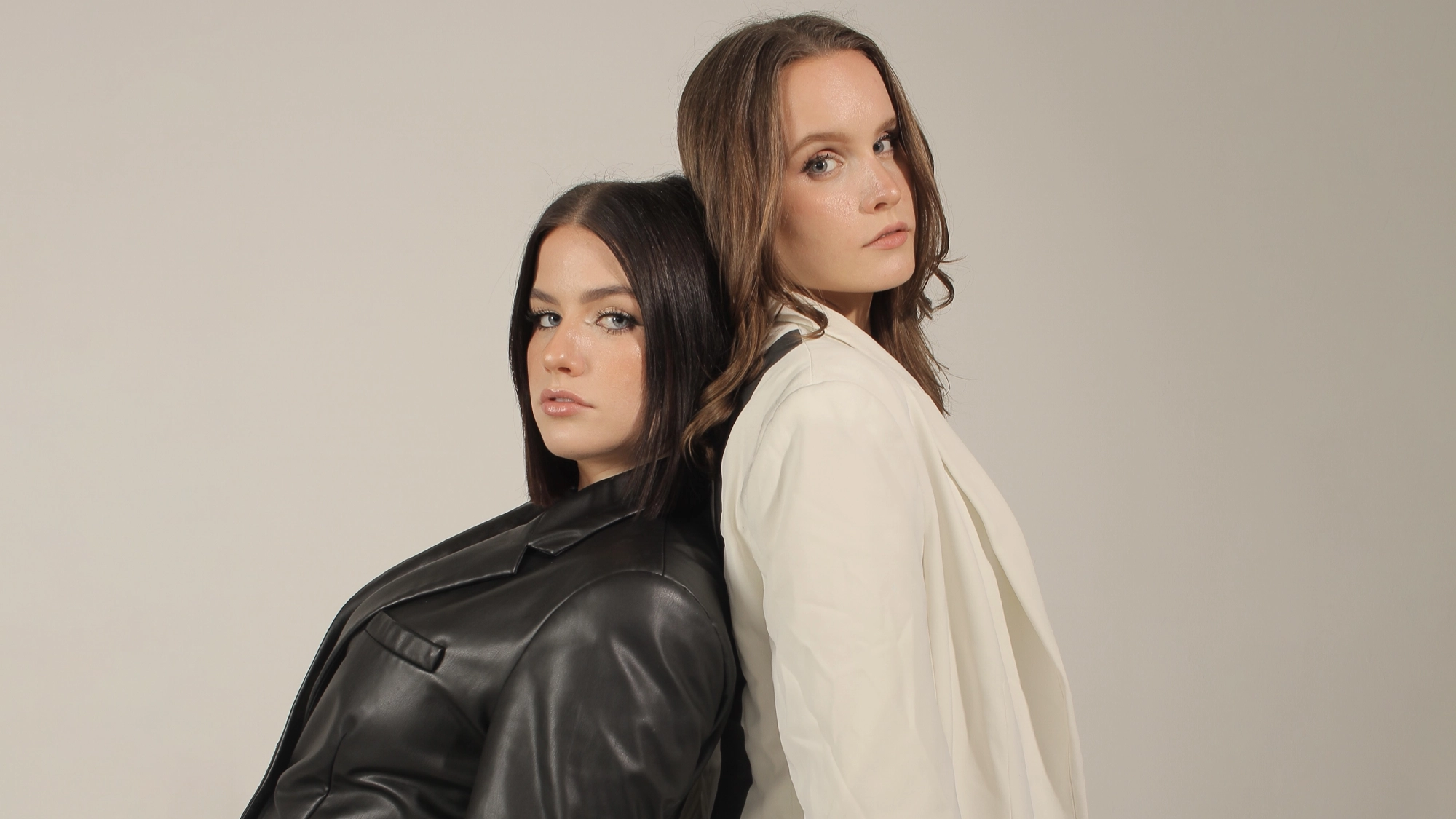 Alexa and Drew Freeman stand back to back against a plain, light gray background. Both look confidently at the camera—Alexa in a black leather jacket, Drew in a white blazer—channeling effortless 