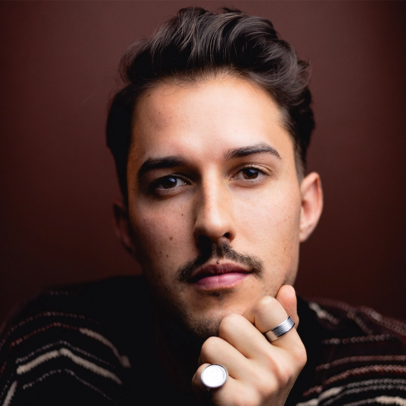 A young man with short, dark hair and a trimmed mustache gazes directly at the camera. He wears a striped sweater and rests his chin on his hand, which displays two silver rings. The background is dark reddish-brown, creating a moody portrait.