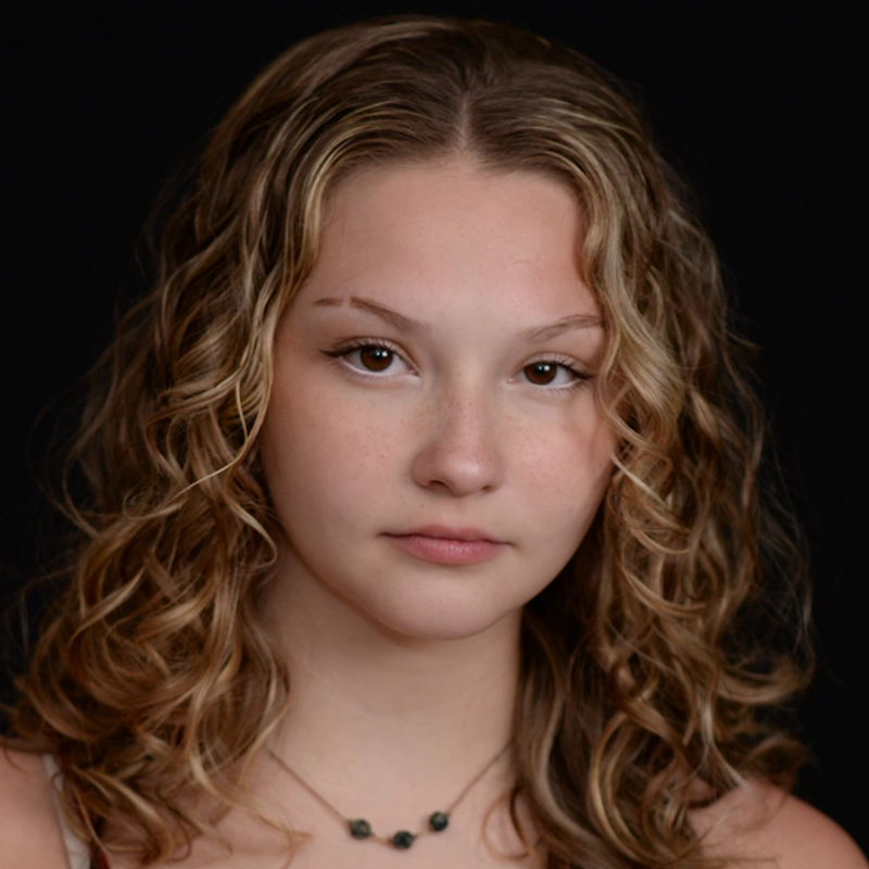 A young woman with light skin and wavy, shoulder-length blonde hair poses against a black background. Addison Zacharias has light brown eyes, subtle makeup, and wears a delicate necklace with small dark beads. Her expression is calm and neutral.