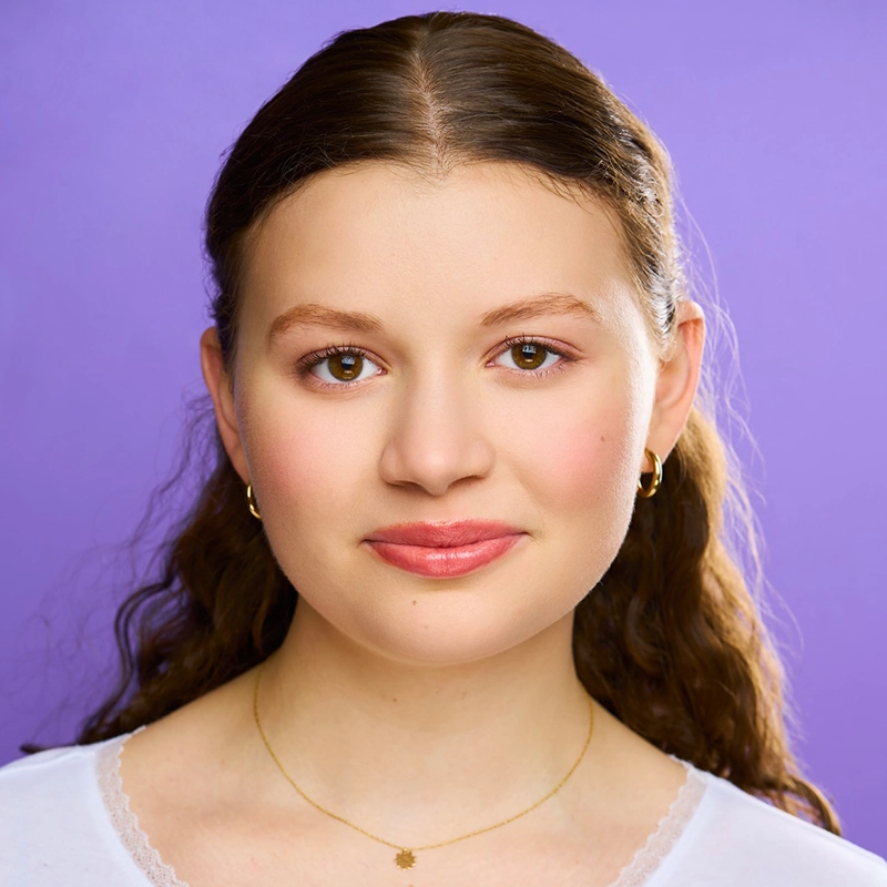 A young woman with wavy brown hair, light skin, and brown eyes smiles softly at the camera. She wears small gold hoop earrings, a delicate gold necklace, and a white top with lace trim. The background is a solid lavender purple.