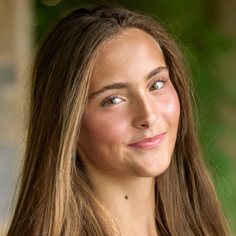 A young woman with long, straight brown hair smiles softly at the camera. She has fair skin, rosy cheeks, and is outdoors with a blurred green background. Micah Wolfensohn wears natural makeup and looks relaxed and confident.