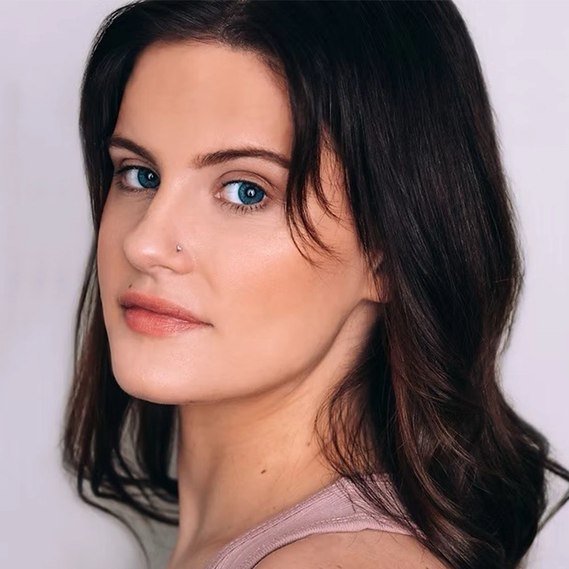 A woman with long brown hair and blue eyes gazes over her shoulder toward the camera. Jordan Whitrock has a small nose stud and wears natural makeup with a light pink top. The soft, neutral background keeps the focus on her face.