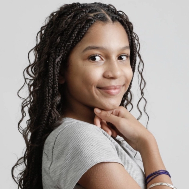 A young girl with long, curly braids and medium-brown skin smiles softly, looking over her shoulder. Anayansi Freya-Williams wears a light gray short-sleeve shirt and two bracelets, her hand gently touching her chin against a plain background.