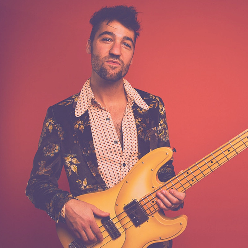 Chris Anderson stands against a red background, holding a yellow electric bass guitar. He wears a black floral jacket over a white shirt with a wide collar and small patterns, and has short, dark hair with a beard, gazing confidently at the camera.
