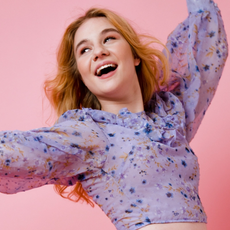 A young woman with long blond hair smiles joyfully, raising her arms. Tracy Lee Anderson wears a light purple, floral-patterned, sheer blouse with puffy sleeves. The solid pink background creates a cheerful, lively mood.