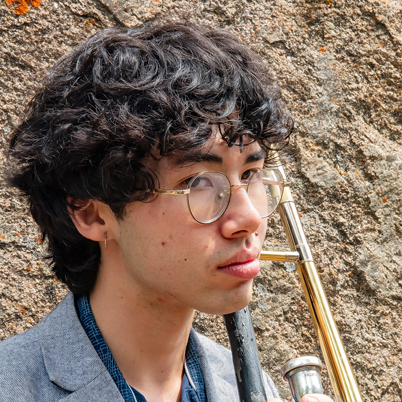 Hannah Mayer, a young person with wavy dark hair, glasses, and an earring, looks to the side while holding a brass trombone. They wear a blue shirt and gray blazer and stand in front of a textured rock wall.