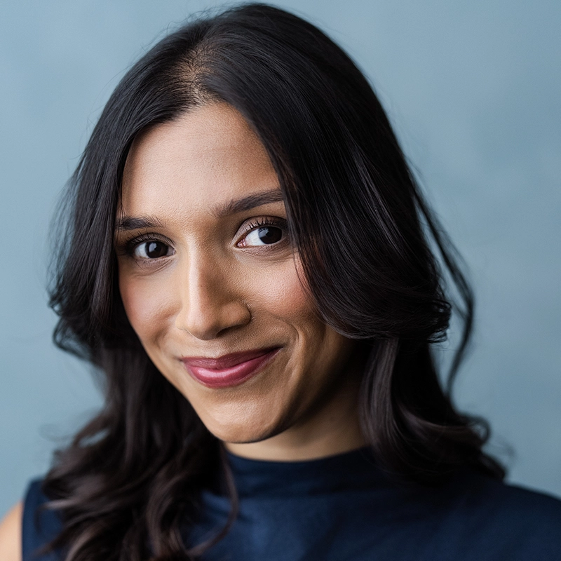 Anika Padwekar, with long, wavy dark hair and medium skin, smiles softly. She wears subtle makeup and a sleeveless navy top. The soft, solid light blue background adds a professional, polished feel to the photo.