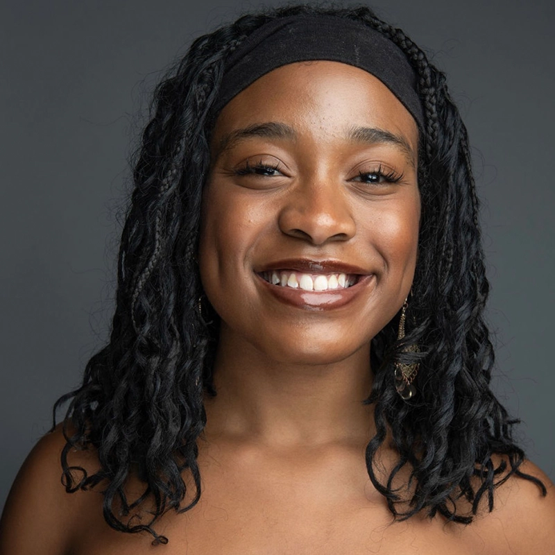 A woman with long, curly black hair wears a black headband and gold dangling earring, smiling brightly. She has brown skin, visible dimples, and is photographed against a plain gray background. Her shoulders are bare.