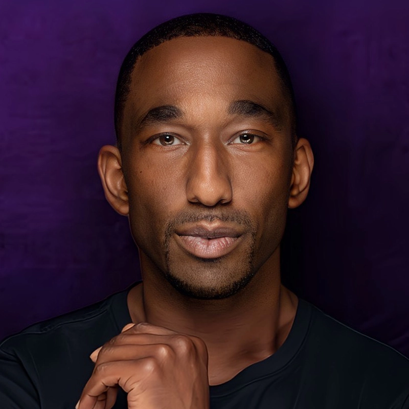 A man with short hair and a trimmed goatee looks confidently at the camera. Channeling the poise of Anthony Wayne, he wears a black shirt against a rich purple background, his hand near his chin in calm self-assurance.