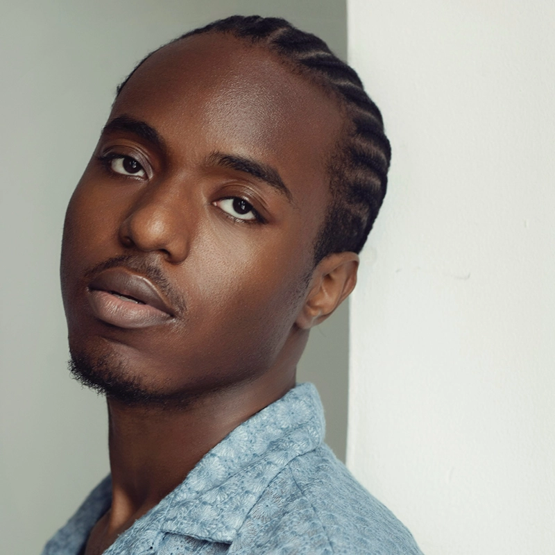 A young Black man with short cornrow braids and a trimmed goatee leans against a white wall. He wears a light blue textured shirt and gazes softly at the camera with a calm, neutral expression. The background is plain and light.