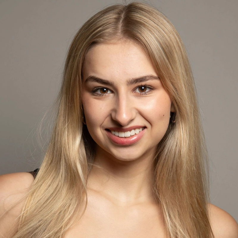 A young woman with long, straight blonde hair smiles at the camera. She has light skin, brown eyes, and is wearing natural makeup. She is dressed in a black top and is posed against a plain, neutral grey background.