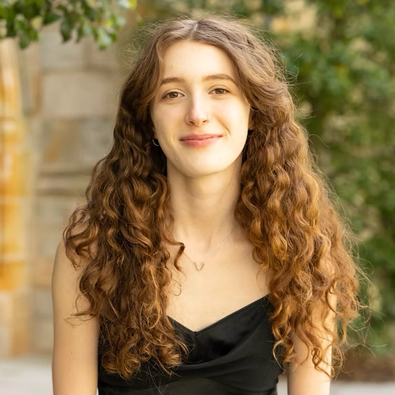 A young woman with long, curly brown hair wears a black sleeveless top and smiles softly at the camera. She is outdoors, with a blurred background of stone and greenery, giving the image a warm, natural feel.