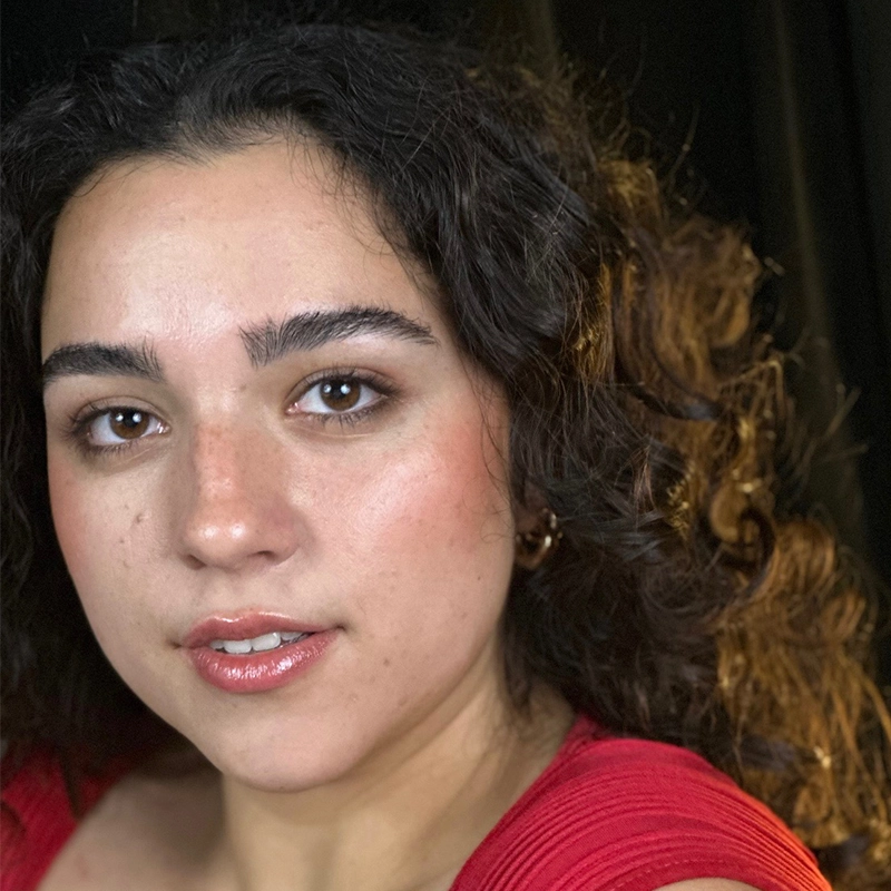 A woman with curly brown hair and thick eyebrows looks at the camera. She has a natural makeup look with glossy lips and wears a red top. The background is dark, highlighting her complexion and the texture of her hair.