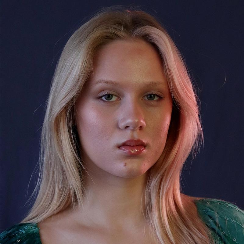 A young woman with long blonde hair and fair skin gazes directly at the camera with a neutral expression. Déa Thatcher wears a green top and glossy lip makeup. The dark background and soft lighting highlight one side of her face and hair.