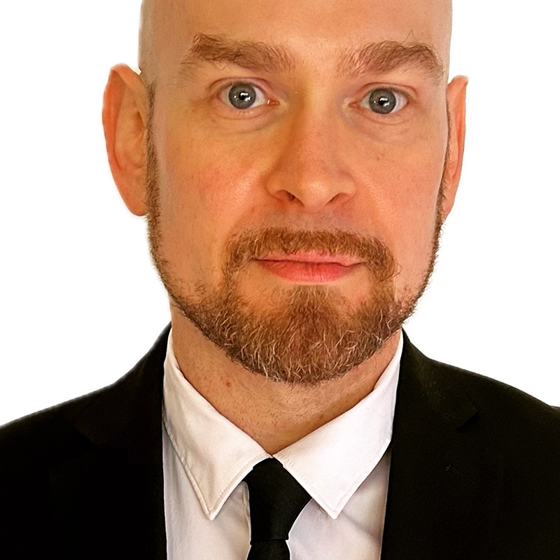 Aron Marchak, a bald man with light skin, blue eyes, and a trimmed beard, is dressed in a black suit, white shirt, and black tie. He looks directly at the camera with a neutral expression against a plain white background.
