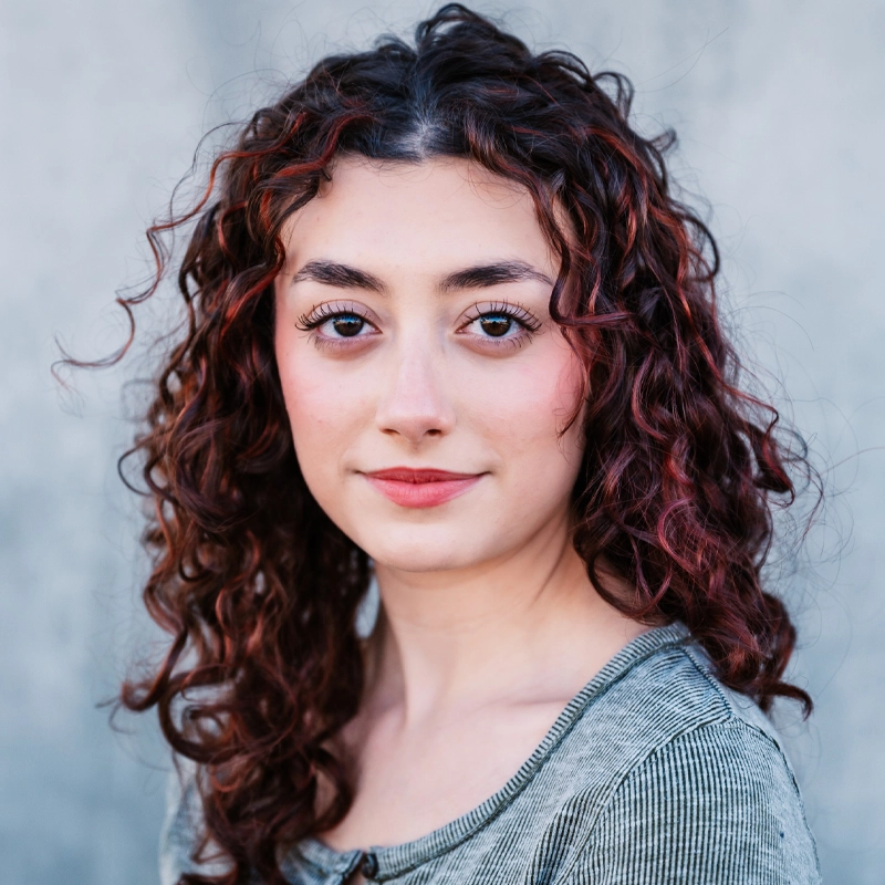 A young woman with fair skin and long, curly brown hair with reddish highlights is facing the camera. She has thick eyebrows, brown eyes, and is wearing a subtle smile. She is dressed in a ribbed, gray-green top with buttons. The background is soft and gray.