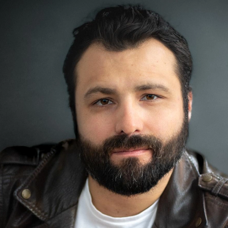 Arturo De La Fuentes, a man with dark hair and a full beard, looks at the camera with a slight smile. He is wearing a brown leather jacket over a white shirt against a plain, dark gray background.