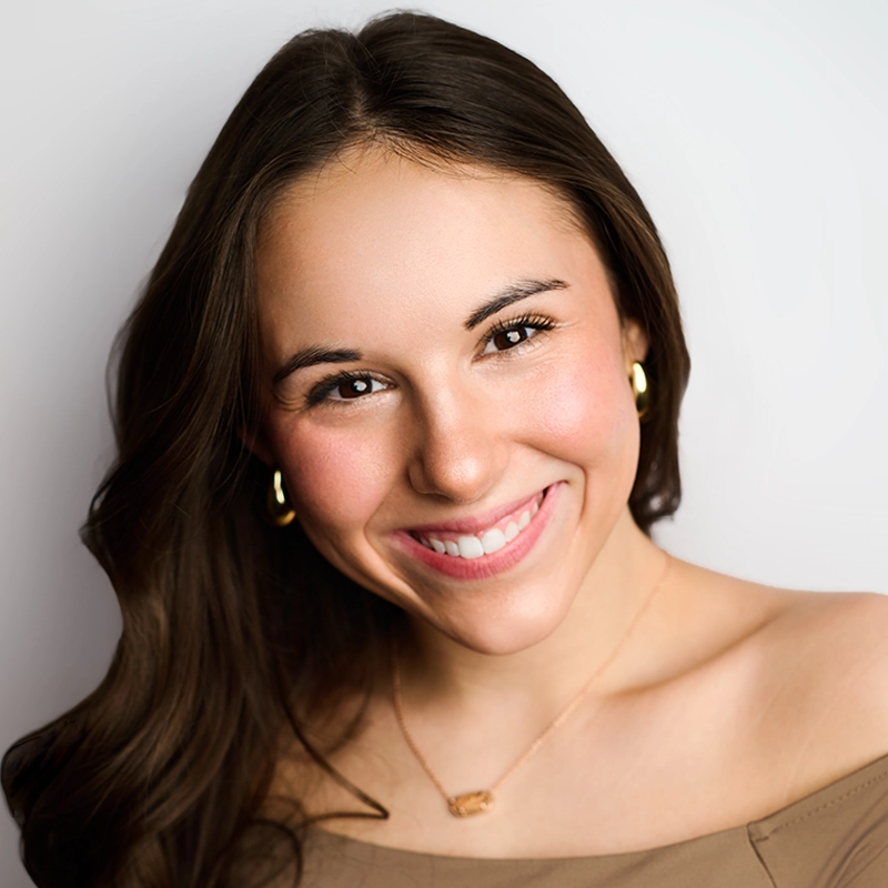 A young woman with long brown hair smiles warmly at the camera. She wears gold hoop earrings, a delicate necklace, and an off-the-shoulder tan top. The background is plain and light, highlighting her friendly expression.